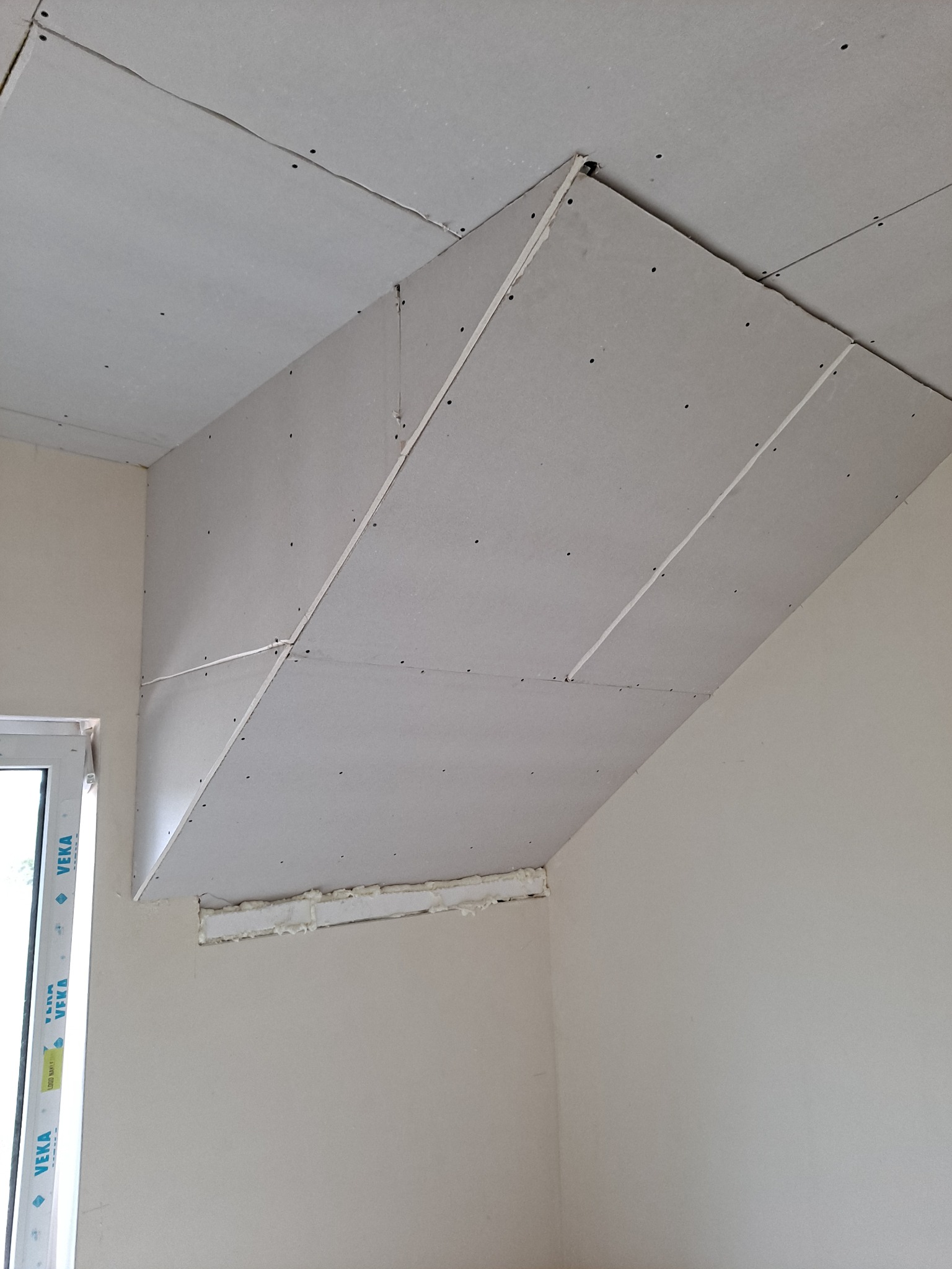 Sufit podwieszany z płyt kartonowo-gipsowych w trakcie montażu, widoczne łączenia i mocowania. Okno z profilem PCV w tle.