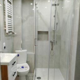 Wykończenia - Nowoczesna łazienka po remoncie: kabina prysznicowa ze szklanymi drzwiami, umywalka z szafką w kolorze drewna i biała toaleta. Szare płytki na ścianach i podłodze.