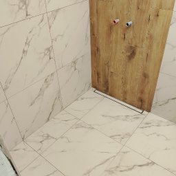 Wykończenia - Wykończona łazienka z białymi płytkami imitującymi marmur na ścianach i podłodze, oraz drewnopodobnymi płytkami w strefie prysznica z odpływem liniowym.