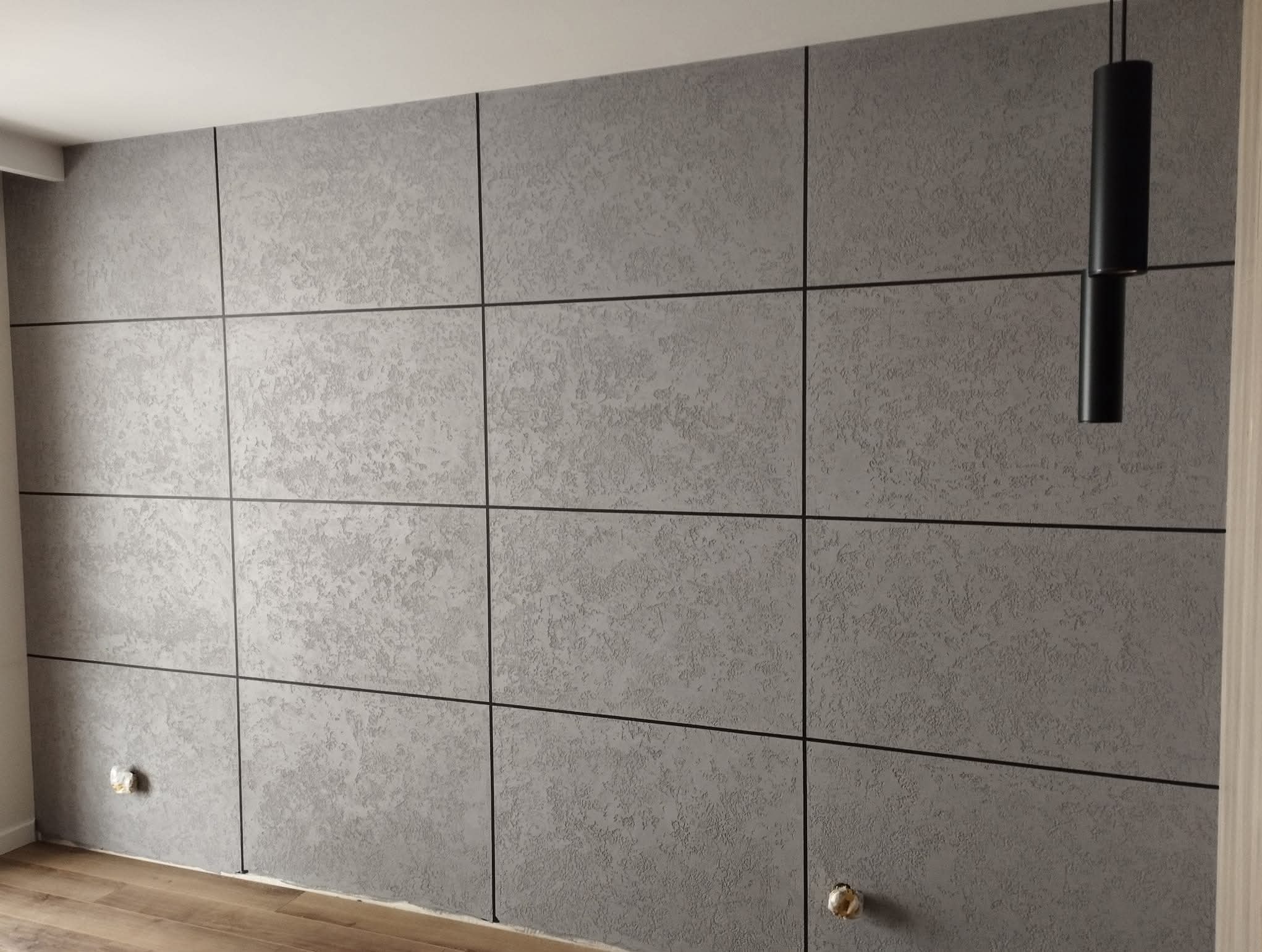 Ściana wykończona szarymi panelami z fakturą, oddzielonymi czarnymi liniami. Widoczna lampa wisząca i fragment podłogi. Nowoczesny design wnętrza.
