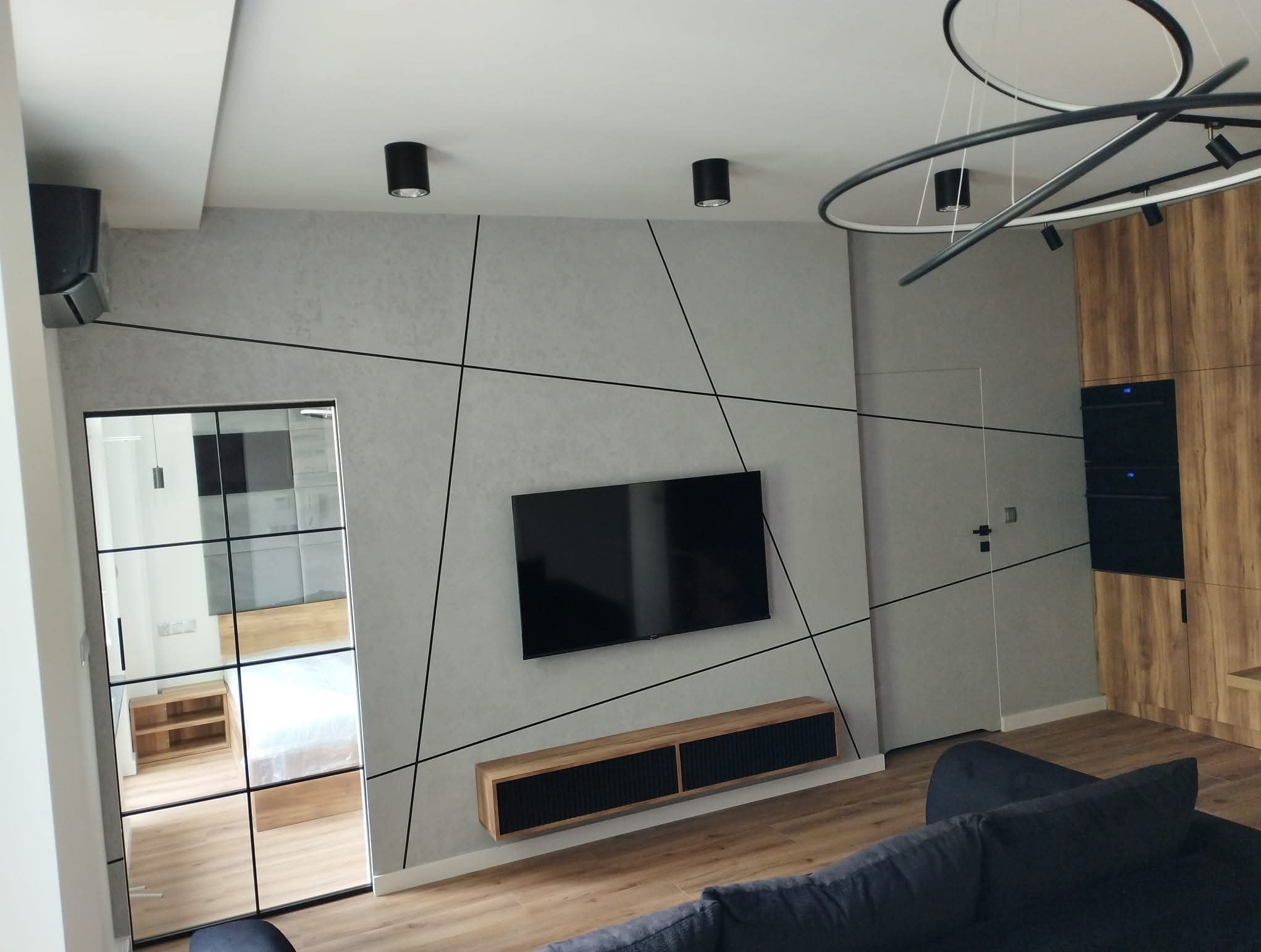 Nowoczesne wykończenie salonu w Dębicy: szara ściana z geometrycznym wzorem, wiszący telewizor i szafka RTV. Widoczne oświetlenie sufitowe i przeszklone drzwi.