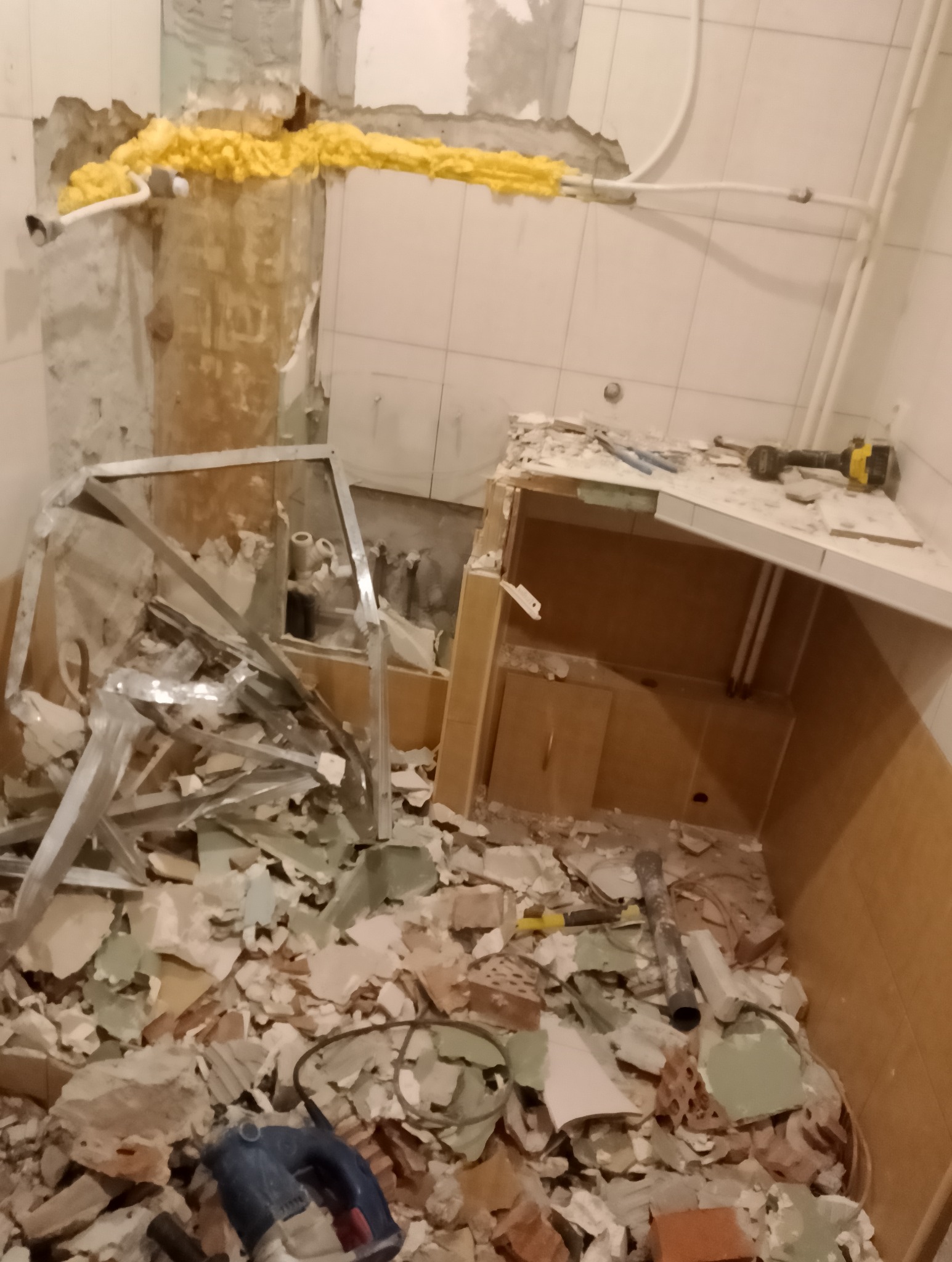 Demontaż starej łazienki: gruz, fragmenty płytek, narzędzia. Widoczne rury zabezpieczone pianką montażową. Etap przygotowania do remontu.
