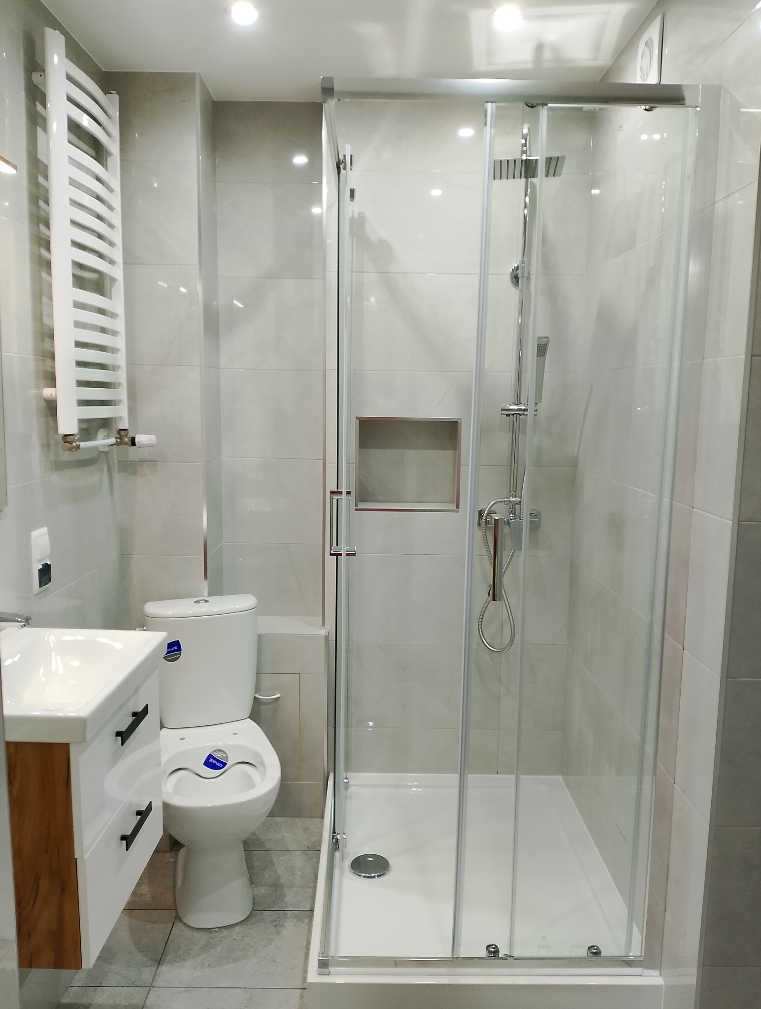 Nowoczesna łazienka po remoncie: kabina prysznicowa ze szklanymi drzwiami, umywalka z szafką w kolorze drewna i biała toaleta. Szare płytki na ścianach i podłodze.