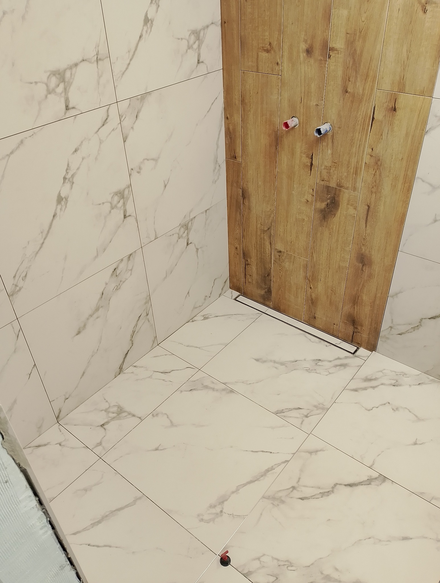 Wykończona łazienka z białymi płytkami imitującymi marmur na ścianach i podłodze, oraz drewnopodobnymi płytkami w strefie prysznica z odpływem liniowym.