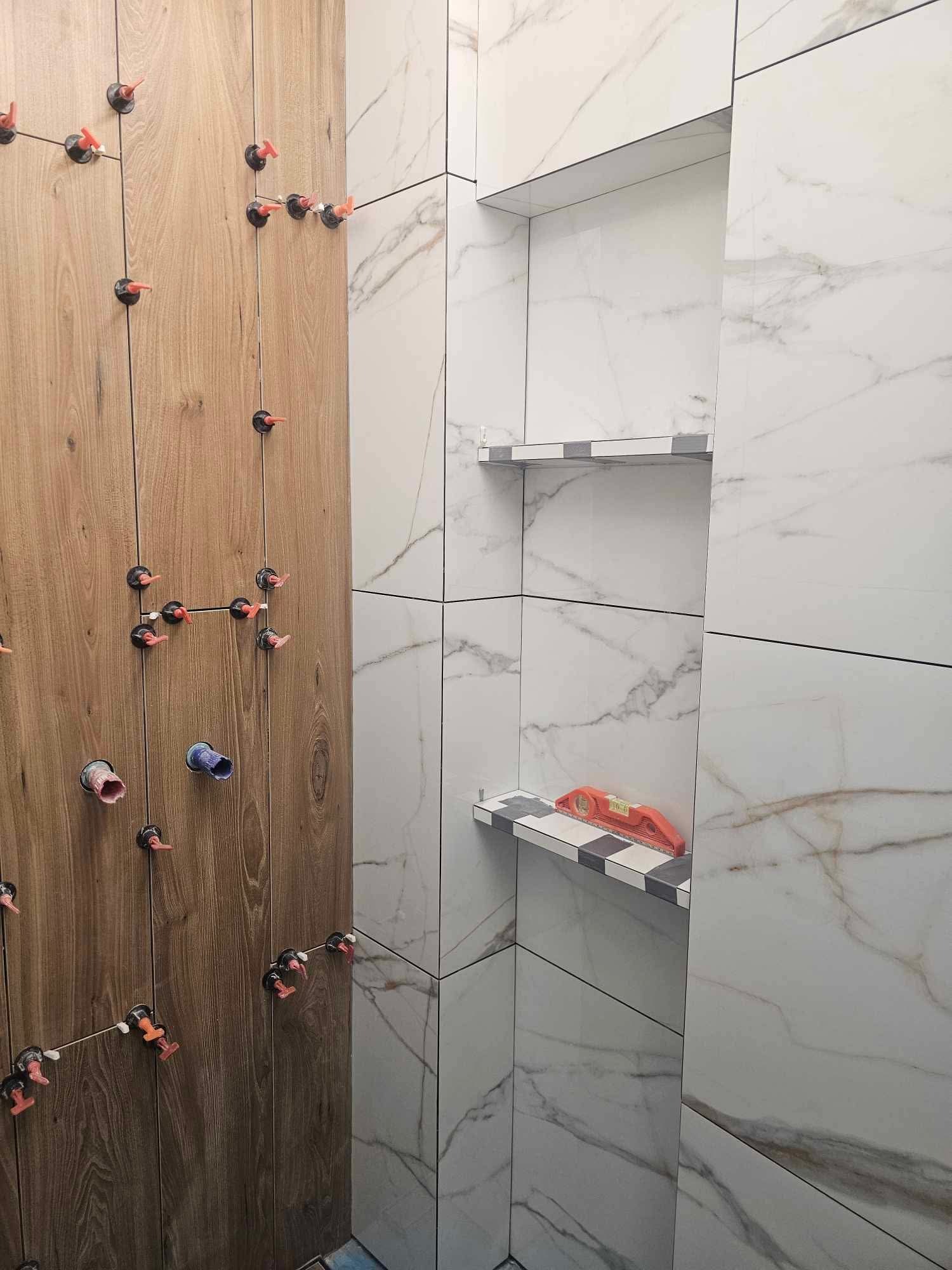 Ściana łazienki w trakcie wykańczania: imitacja drewna i marmurowe płytki, system poziomowania płytek, półki wnękowe z poziomnicą. Detal układania płytek.