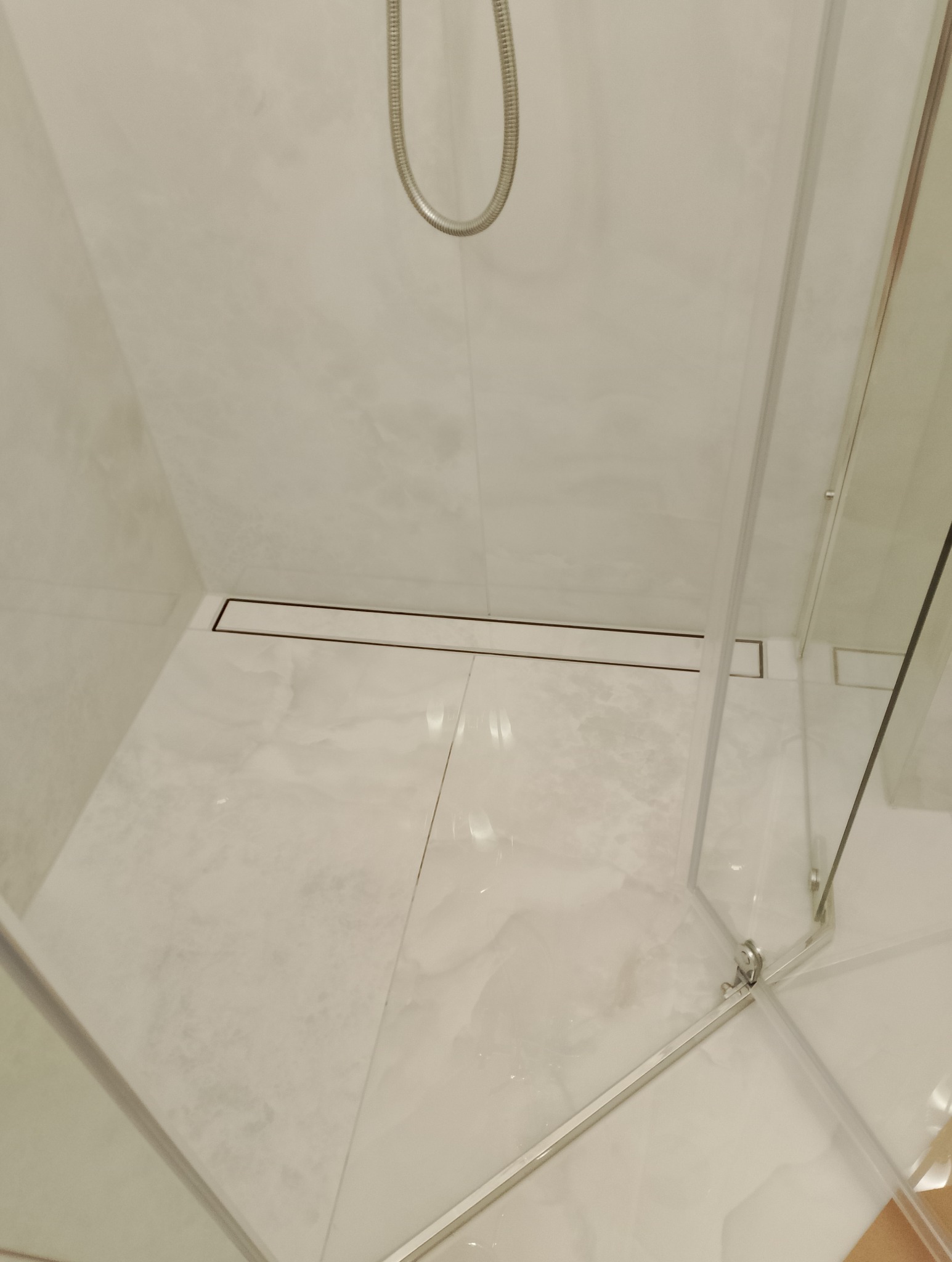 Biała kabina prysznicowa z odpływem liniowym i marmurowymi płytkami. Widok z góry, minimalistyczny design, chromowane wykończenia.