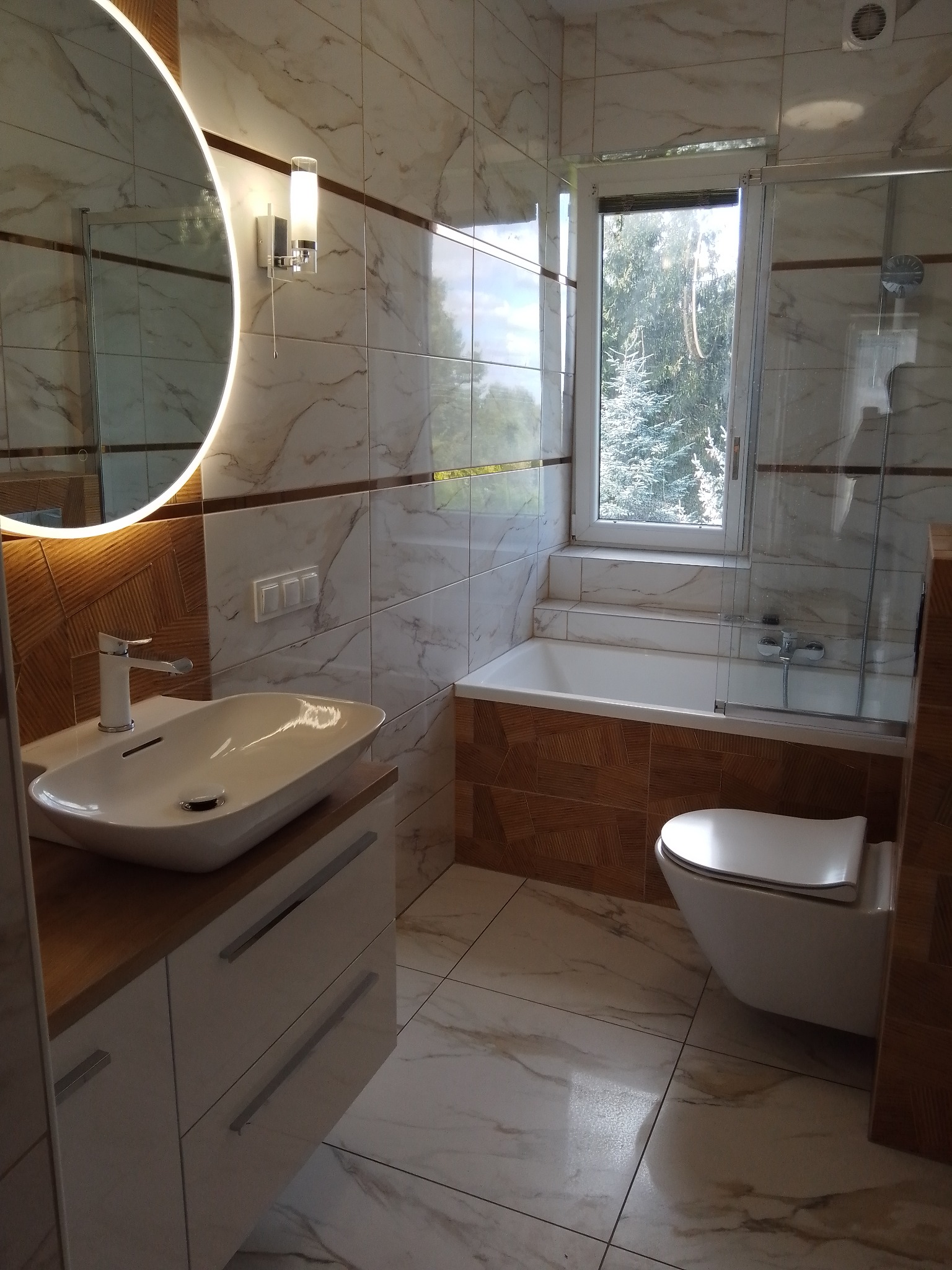 Jasna łazienka z marmurowymi płytkami, okrągłym lustrem z podświetleniem LED, białą umywalką nablatową i podwieszaną toaletą. Widok na okno z zielenią.