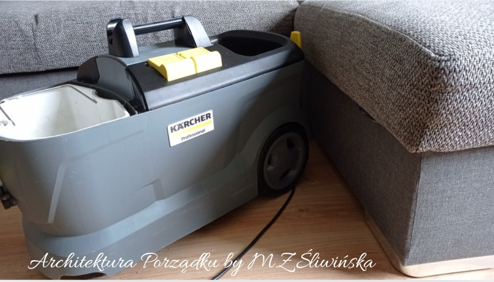 Profesjonalny odkurzacz piorący Kärcher obok szarej sofy. Widoczny kabel zasilający. Sprzęt do prania tapicerki w trakcie pracy.