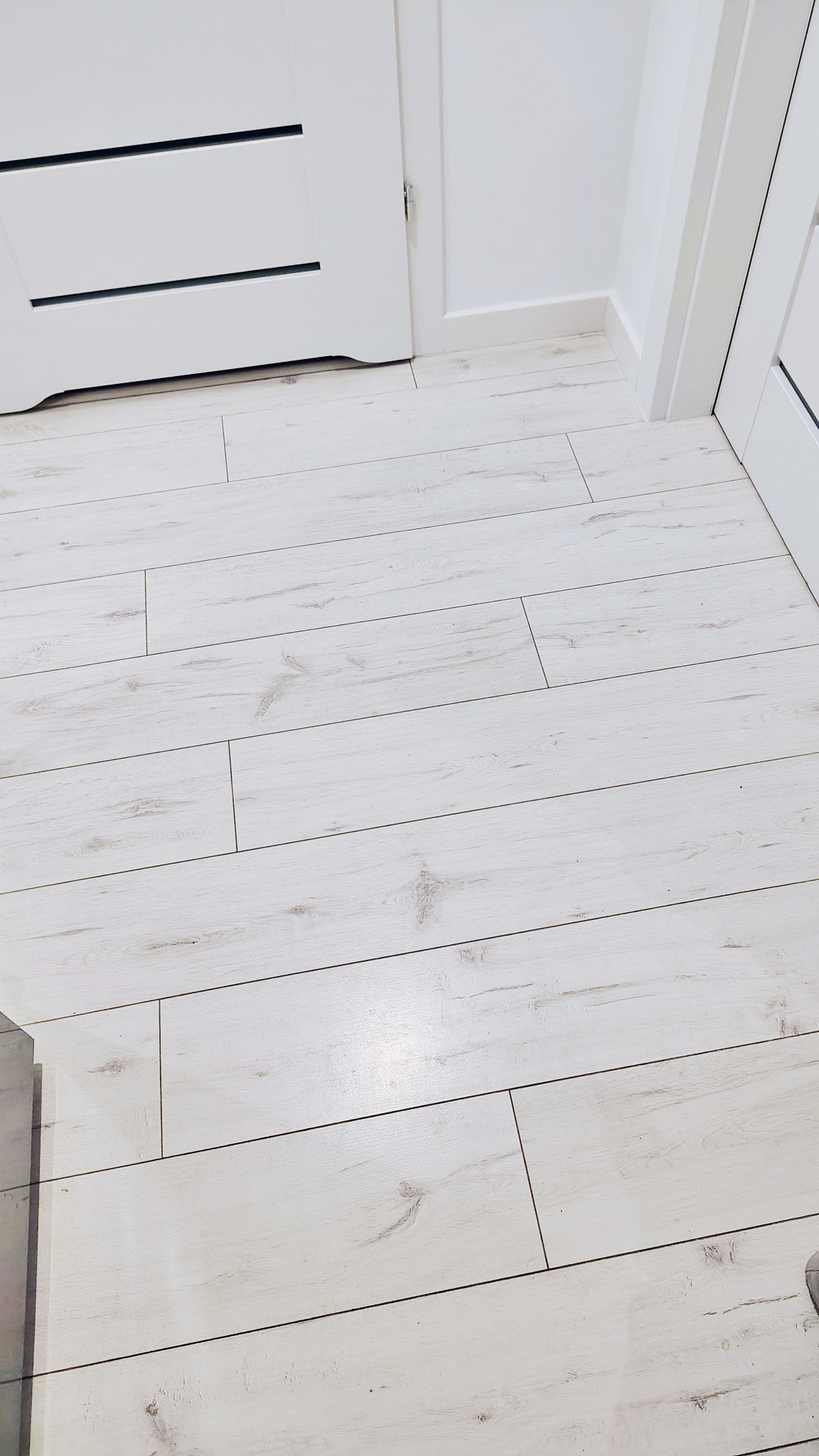 Jasne, drewnopodobne panele podłogowe ułożone wzdłuż ściany z białymi drzwiami i listwami przypodłogowymi. Minimalistyczny design, czyste linie, skandynawski styl.