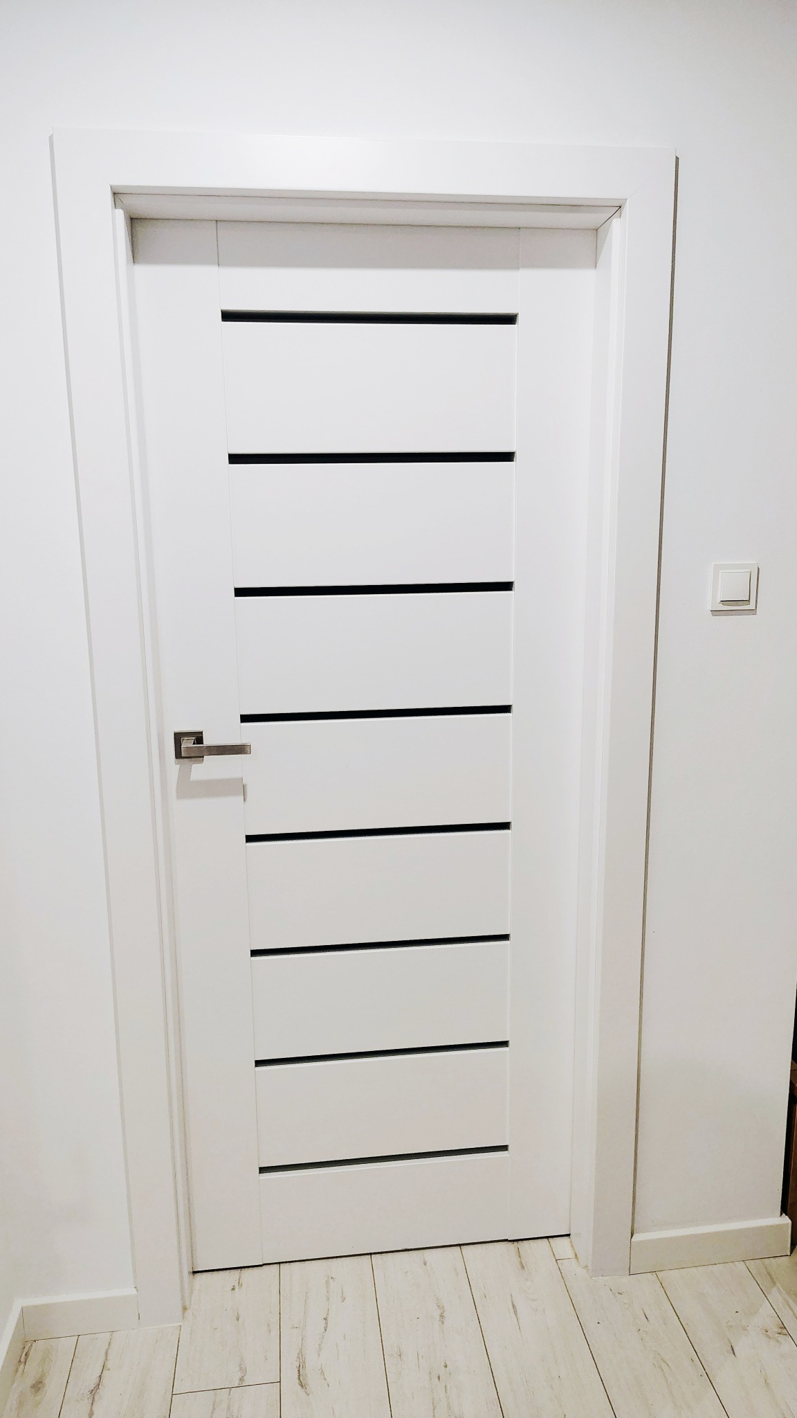 Białe, nowoczesne drzwi z poziomymi, czarnymi wstawkami i srebrną klamką, zamontowane w białej ścianie z włącznikiem. Jasna podłoga z paneli.