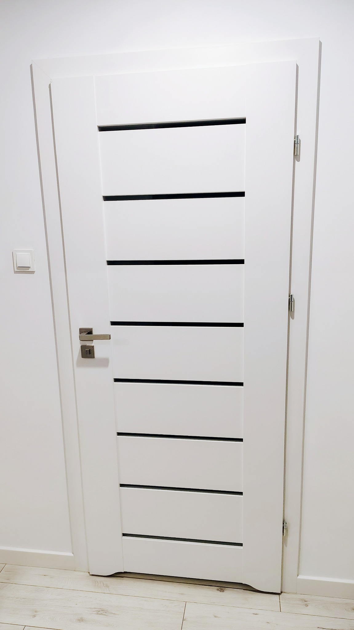 Białe drzwi wewnętrzne z poziomymi, czarnymi wstawkami, zamontowane w białej ścianie. Widoczny fragment podłogi z jasnymi panelami i włącznik światła.