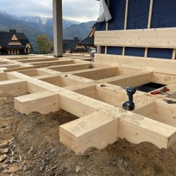 USŁUGI REMONTOWO WYKOŃCZENIOWE BUDOWNICTWO KONSTRUKCYJNE - Drewniana konstrukcja stropu z widocznymi belkami i narzędziami na tle górskiego krajobrazu. Elementy wykończeniowe w stylu zakopiańskim.