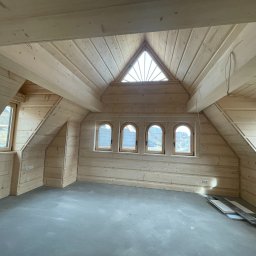 USŁUGI REMONTOWO WYKOŃCZENIOWE BUDOWNICTWO KONSTRUKCYJNE - Drewniane wykończenie poddasza z oknami o różnych kształtach: trójkątnym i łukowym. Widoczne deski podłogowe i gniazdka elektryczne na ścianie.