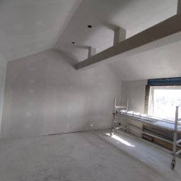 USŁUGI REMONTOWO WYKOŃCZENIOWE BUDOWNICTWO KONSTRUKCYJNE - Wnętrze poddasza po szpachlowaniu, z widocznymi elementami konstrukcyjnymi i rusztowaniem. Ściany i sufit w jasnym kolorze, przygotowane do dalszych prac wykończeniowych.