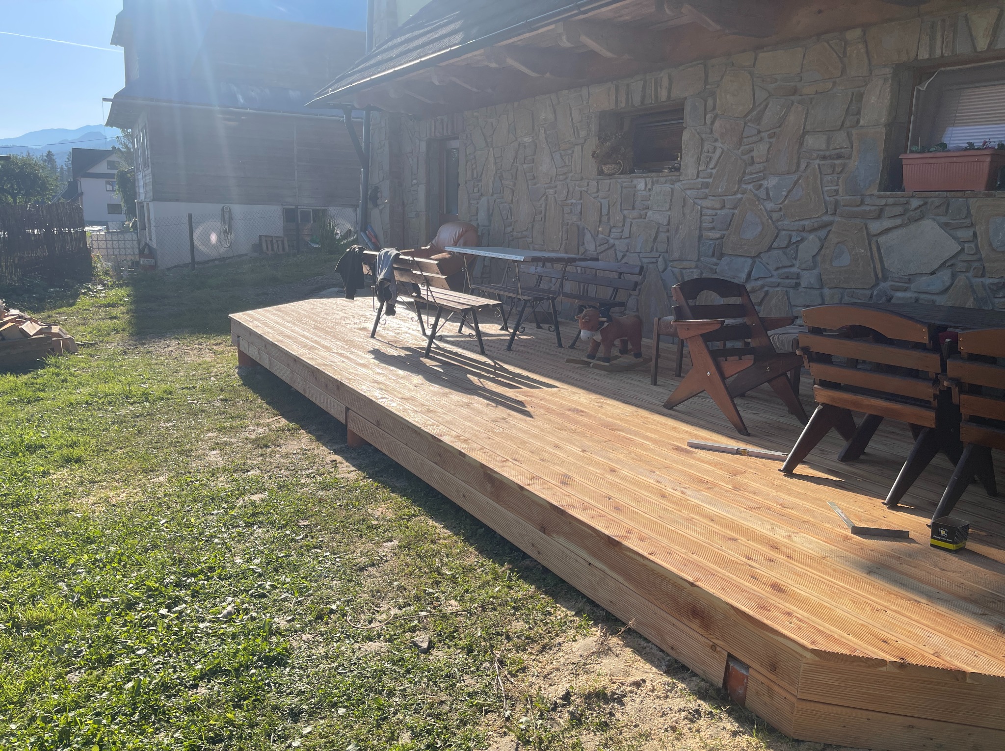 Nowy drewniany taras przy kamiennym domu w słoneczny dzień. Widoczne narzędzia i meble ogrodowe. Tło stanowią góry i zieleń. Jasne, naturalne światło.