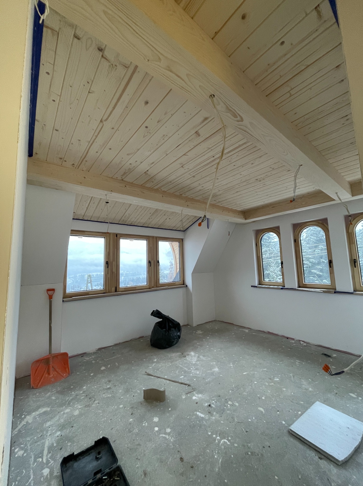 Surowe wnętrze poddasza z drewnianą boazerią sufitową, okna łukowe i narzędzia budowlane na podłodze. Widok na góry za oknem.