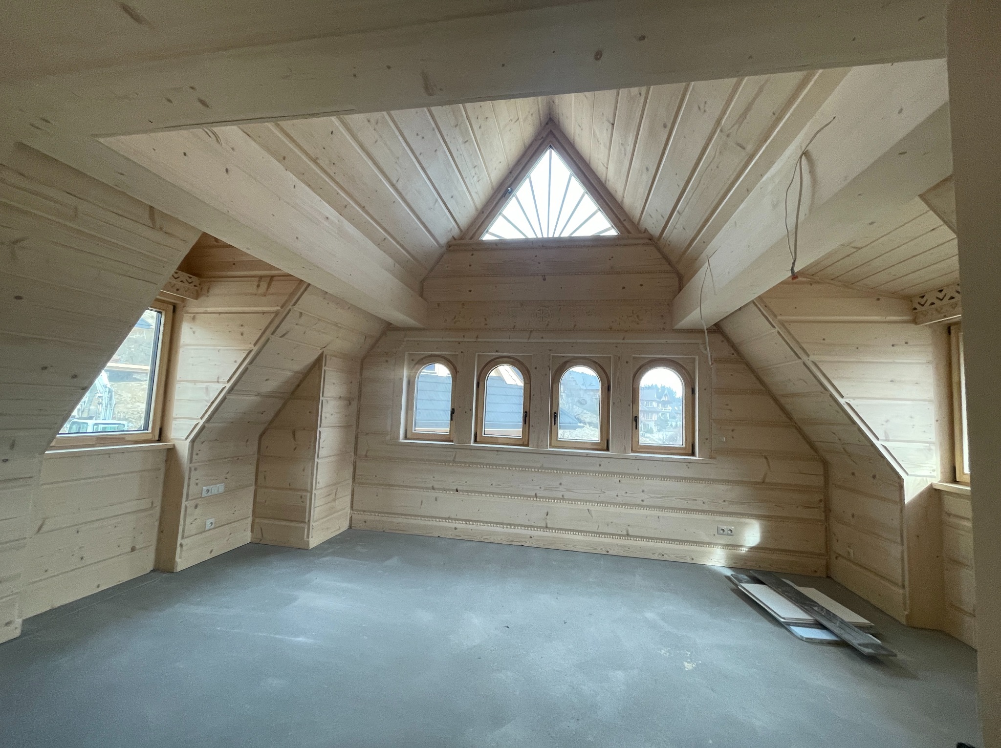 Drewniane wykończenie poddasza z oknami o różnych kształtach: trójkątnym i łukowym. Widoczne deski podłogowe i gniazdka elektryczne na ścianie.