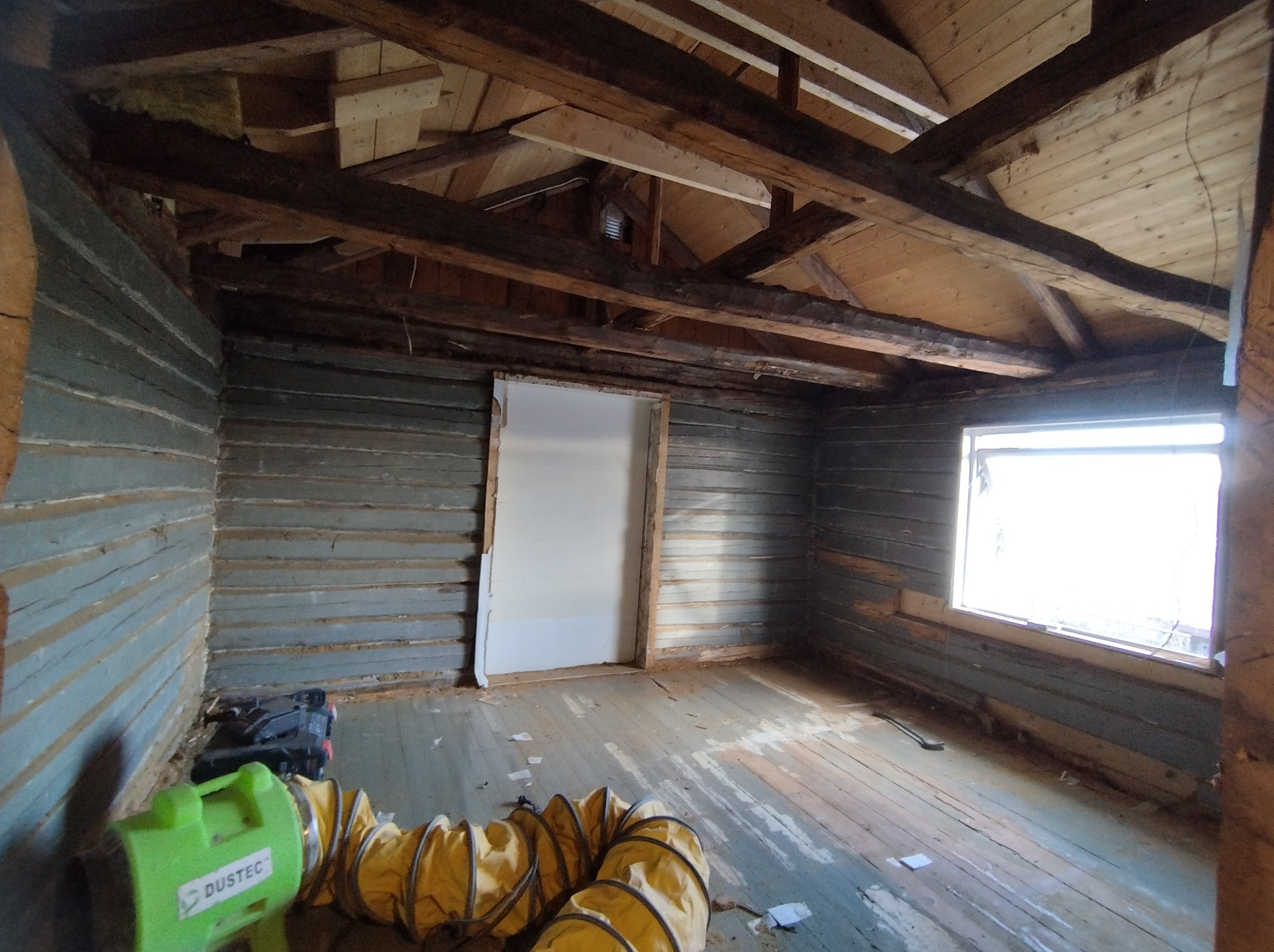 Wnętrze remontowanego drewnianego domu z odsłoniętą konstrukcją dachu, widoczne narzędzia i wentylator. Ściany pomalowane na niebiesko, okno z jasnym światłem.