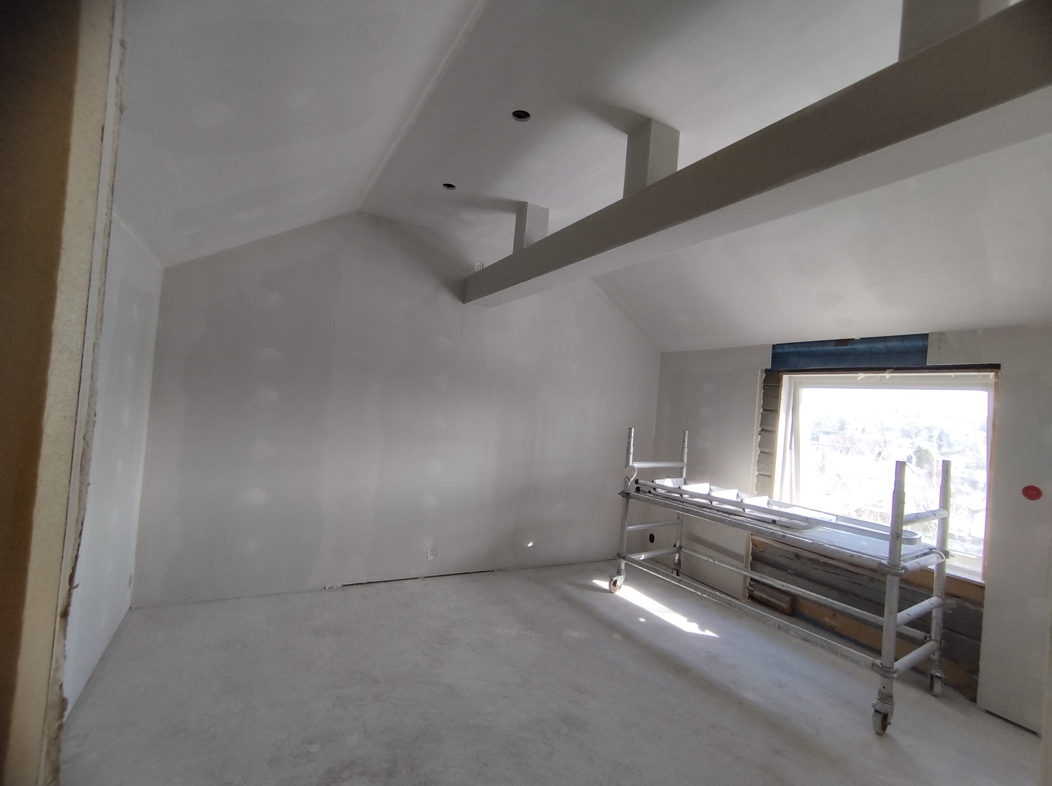 Wnętrze poddasza po szpachlowaniu, z widocznymi elementami konstrukcyjnymi i rusztowaniem. Ściany i sufit w jasnym kolorze, przygotowane do dalszych prac wykończeniowych.