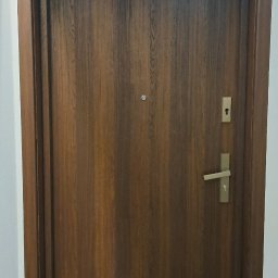 TWINCONCEPT - Zamknięte, brązowe drzwi wejściowe z drewnianą ramą, widoczne od frontu, z zamontowaną klamką i wizjerem. Pod drzwiami pomarańczowy próg zabezpieczający.