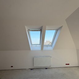 TWINCONCEPT - Białe poddasze z dwoma oknami dachowymi, grzejnikiem i narzędziami malarskimi na podłodze. Widok na błękitne niebo i fragment dachu sąsiedniego budynku.