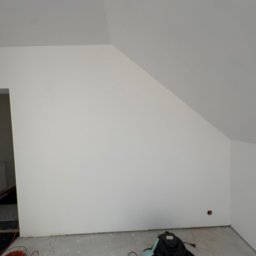 TWINCONCEPT - Świeżo pomalowane na biało poddasze ze skosami. Na podłodze wiadro z farbą, odkurzacz i zwinięty kabel. Widoczny fragment drzwi i grzejnika.