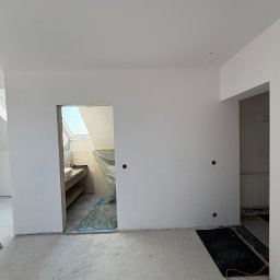 TWINCONCEPT - Wnętrze w trakcie prac wykończeniowych: białe ściany, surowa podłoga, widoczne instalacje elektryczne i fragment łazienki z oknem dachowym w tle.