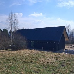 TWINCONCEPT - Nowoczesny dom modułowy z ciemną elewacją i dwuspadowym dachem, otoczony naturalnym krajobrazem z drzewami i łąką. Widoczne materiały budowlane obok domu.