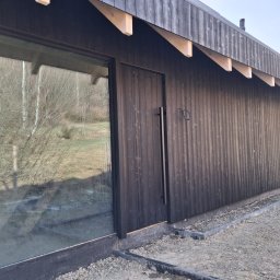 TWINCONCEPT - Nowoczesny dom modułowy z ciemną, drewnianą elewacją i dużym oknem, otoczony żwirem. Widoczne elementy konstrukcyjne dachu i minimalistyczne drzwi.