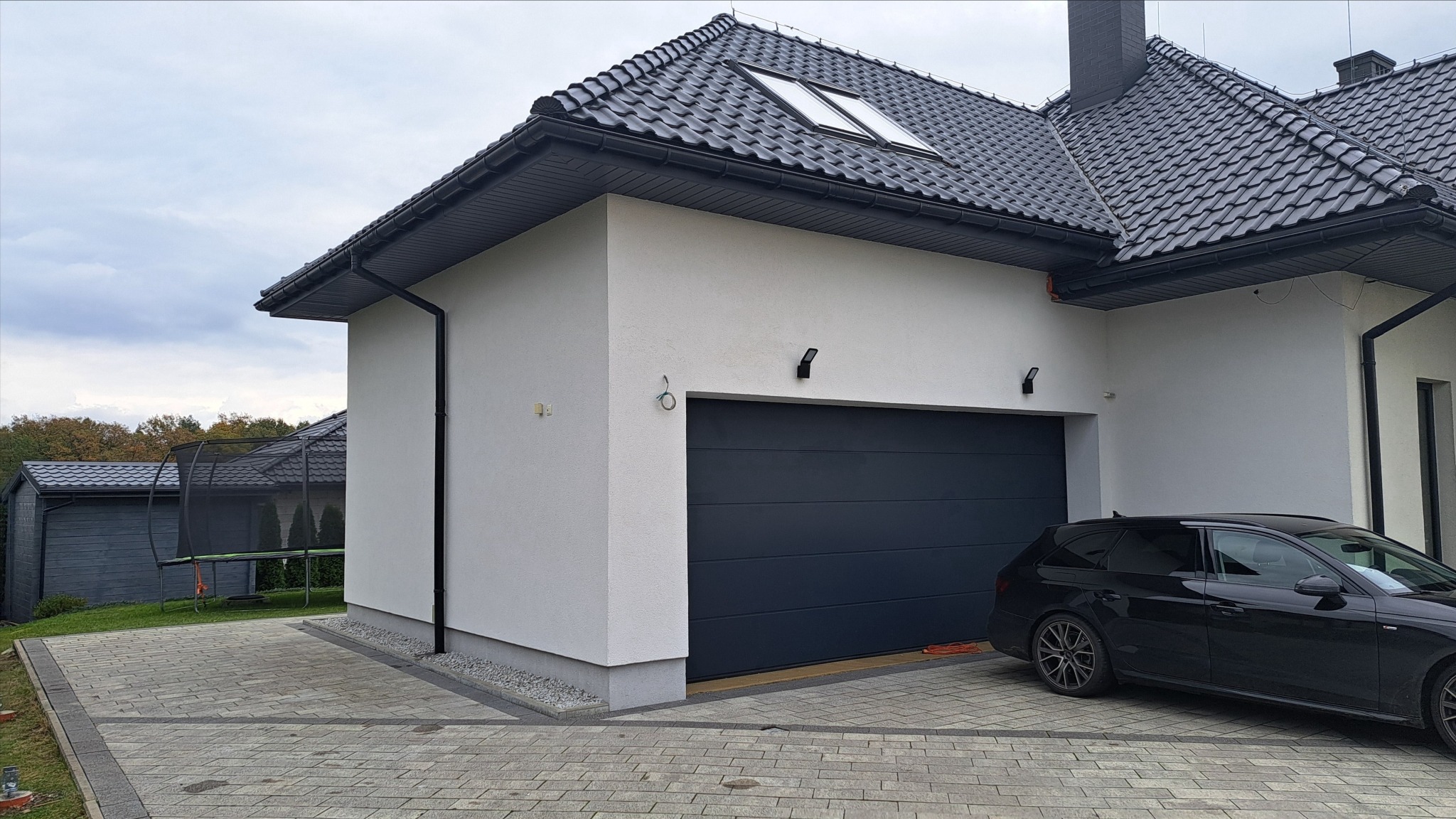 Biała elewacja domu z grafitową bramą garażową i czarnym dachem. Widoczny fragment czarnego samochodu. Kostka brukowa na podjeździe. Nowoczesny styl.