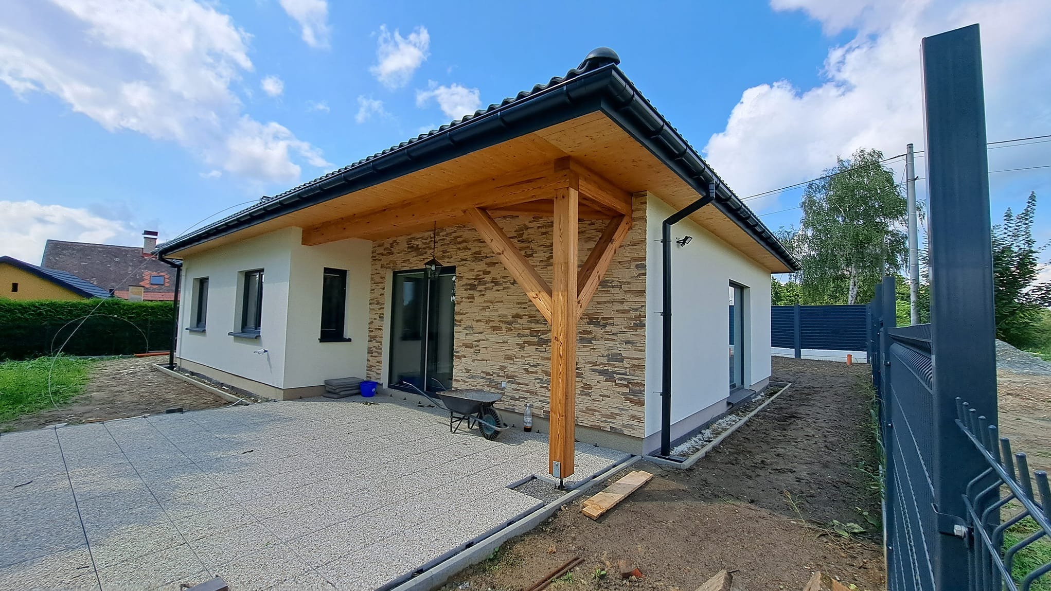 Nowoczesny dom jednorodzinny po remoncie elewacji z drewnianą pergolą i wykończonym tarasem z kostki brukowej. Widok z boku, słoneczny dzień.