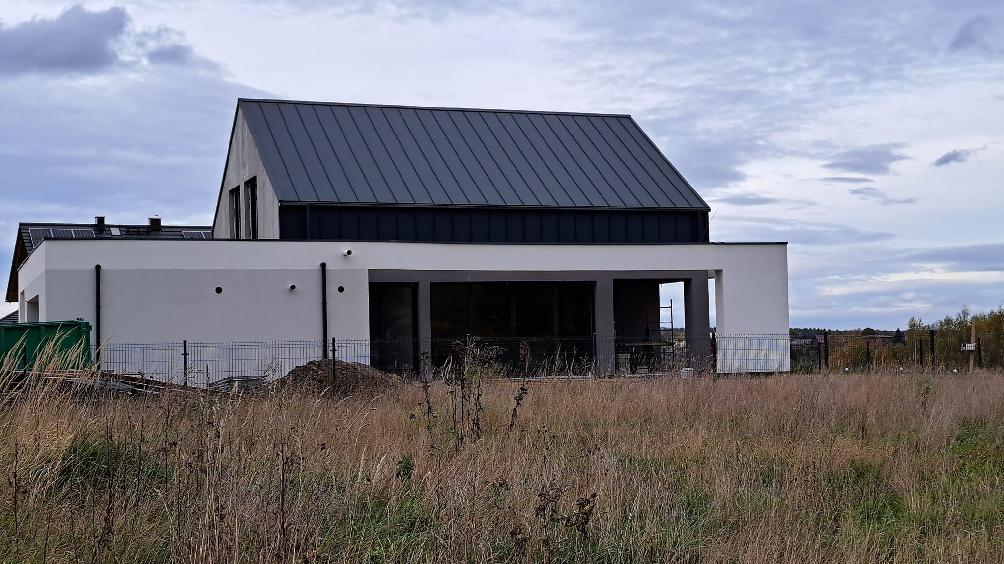 Nowoczesny dom z szarą, metalową elewacją dachu i białą elewacją ścian, otoczony wysoką trawą i ogrodzeniem z siatki. Widoczne elementy rusztowania.