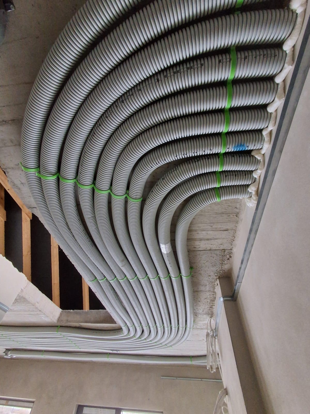 Szereg szarych, elastycznych rur rekuperacji, przymocowanych zielonymi opaskami do sufitu. Widoczne elementy konstrukcyjne budynku i pianka uszczelniająca.