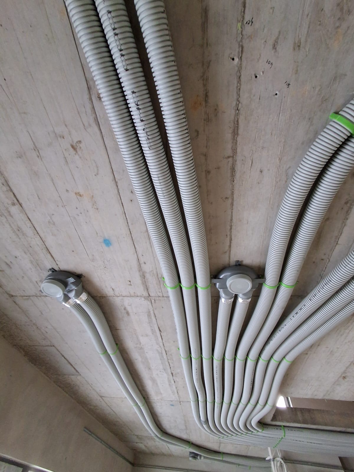 Szare, karbowane rury wentylacyjne przymocowane do surowego betonowego sufitu za pomocą zielonych opasek zaciskowych i szarych puszek instalacyjnych.