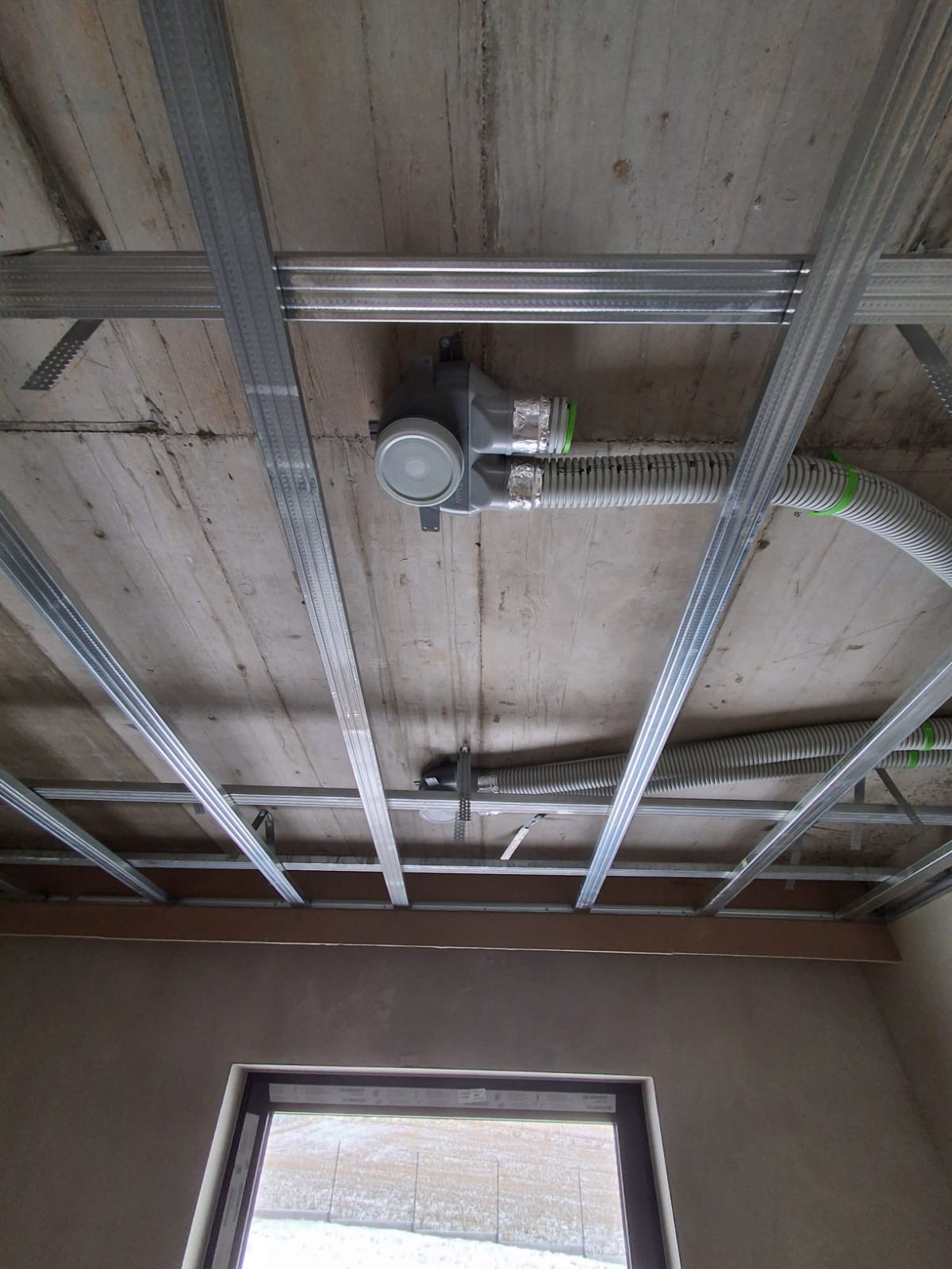 Montaż instalacji rekuperacji pod stropem z widocznymi przewodami wentylacyjnymi i elementami mocującymi. Widok z dołu na sufit w trakcie prac instalacyjnych.
