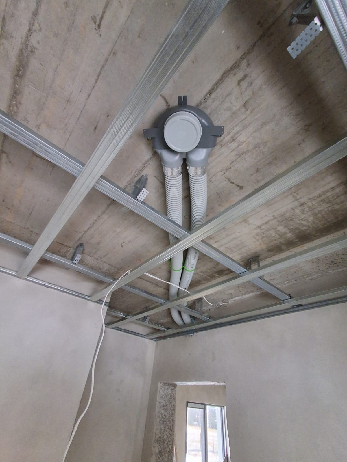 Szara centrala rekuperacyjna z dwoma elastycznymi przewodami wentylacyjnymi, zamontowana pod surowym stropem z metalową konstrukcją nośną.