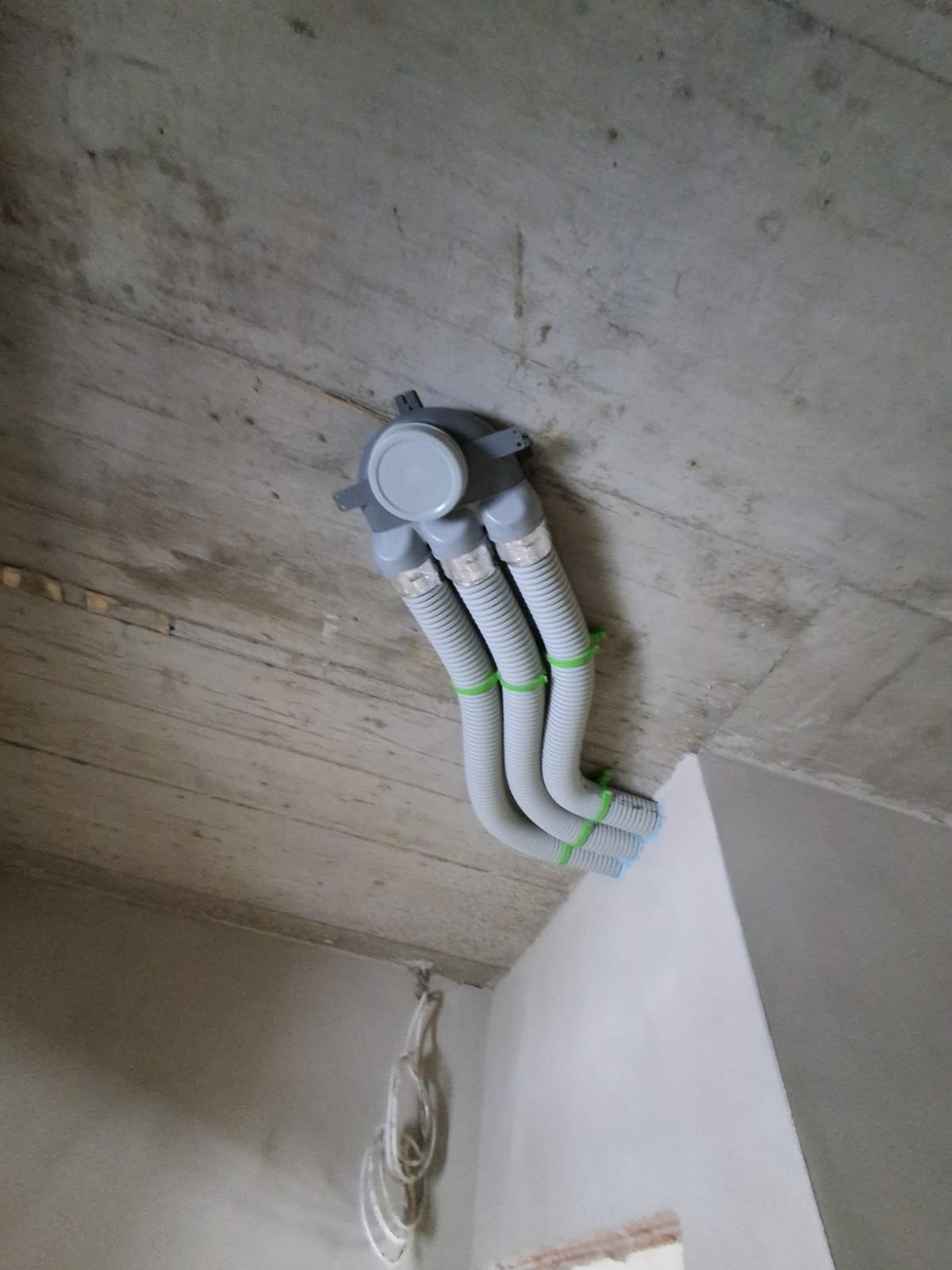 Szare, elastyczne rury wentylacyjne z zielonymi opaskami przymocowane do surowego, betonowego sufitu w rogu pomieszczenia. Widoczne kable na ścianie.