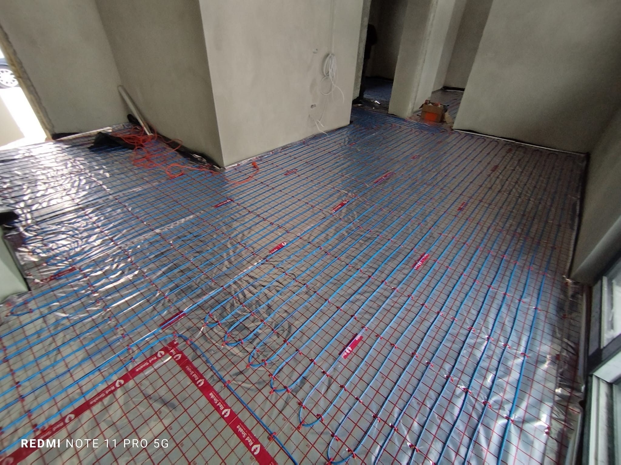 Rozłożona instalacja ogrzewania podłogowego z niebieskimi rurkami na folii aluminiowej w surowym wnętrzu. Widoczne czerwone elementy mocujące rurki do siatki.