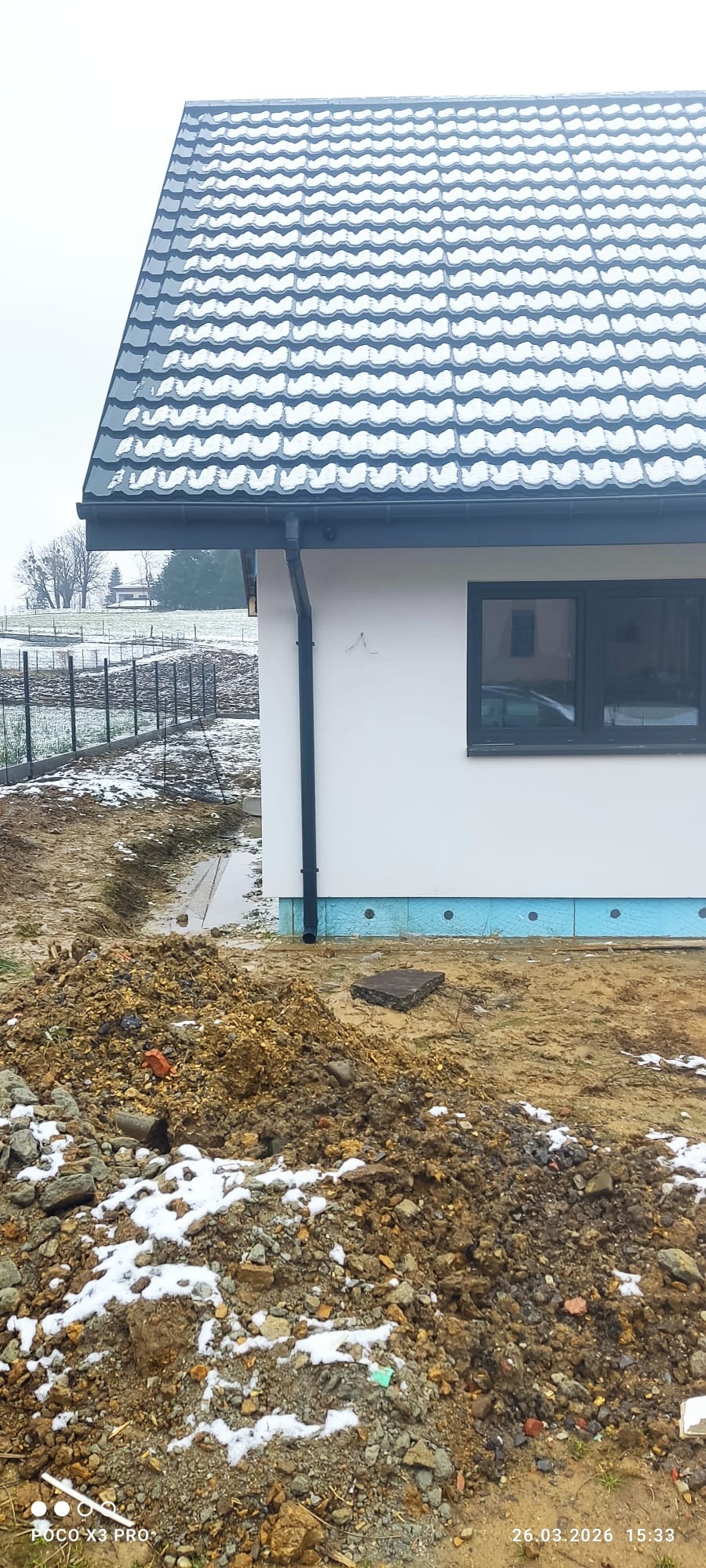Fragment domu z białą elewacją, czarną rynną i oknem. Widoczny fragment dachu z szarą dachówką. Na pierwszym planie ziemia z resztkami śniegu.