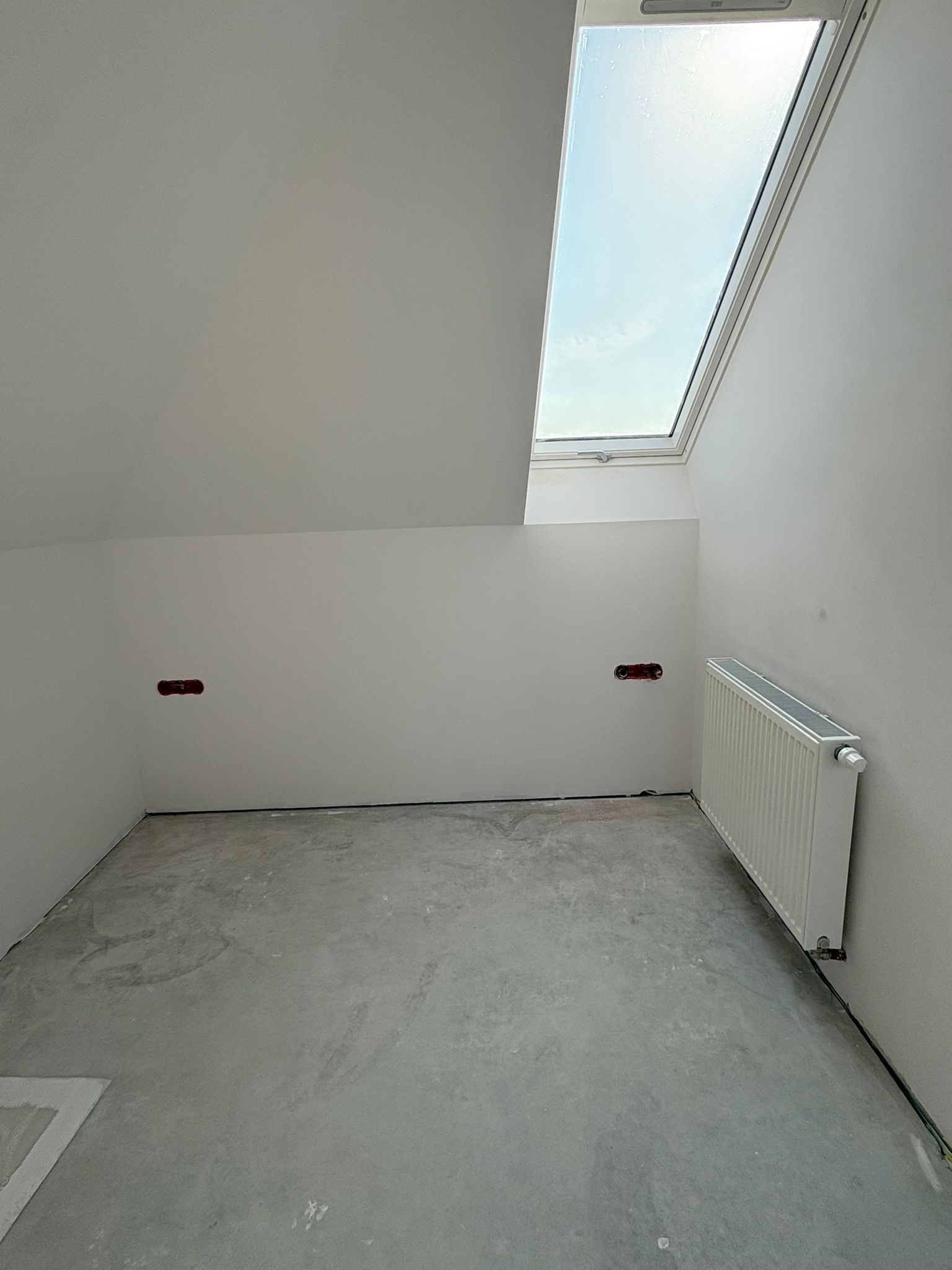 Wnętrze poddasza z oknem dachowym, grzejnikiem i przygotowanymi punktami elektrycznymi. Ściany pomalowane na biało, surowa podłoga. Widok na niebo przez okno.