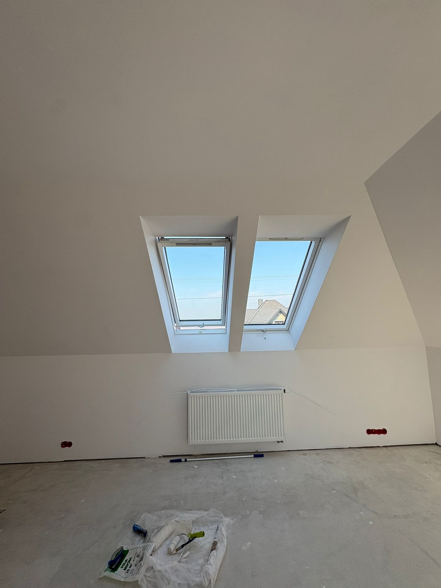 Białe poddasze z dwoma oknami dachowymi, grzejnikiem i narzędziami malarskimi na podłodze. Widok na błękitne niebo i fragment dachu sąsiedniego budynku.