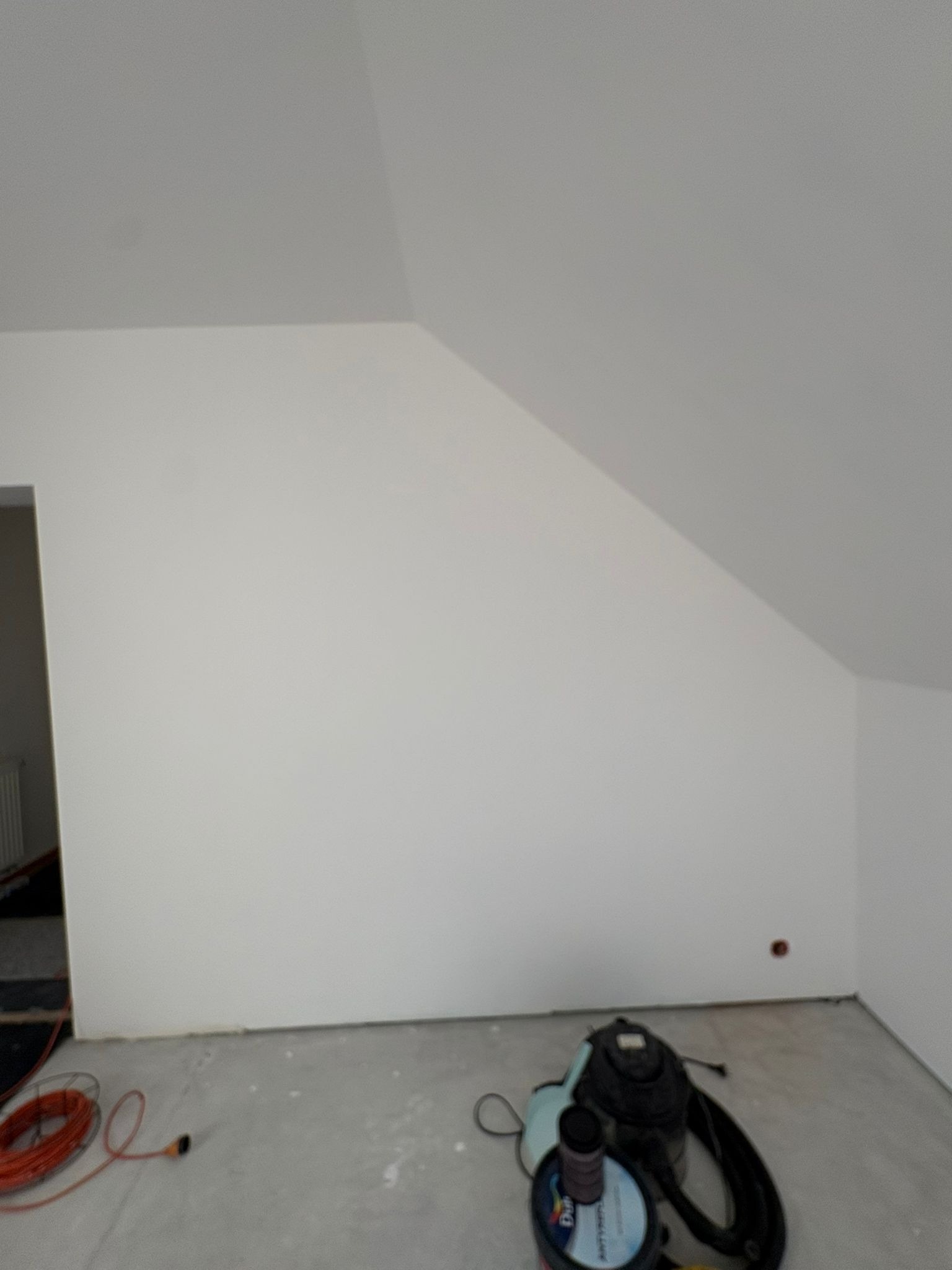 Świeżo pomalowane na biało poddasze ze skosami. Na podłodze wiadro z farbą, odkurzacz i zwinięty kabel. Widoczny fragment drzwi i grzejnika.