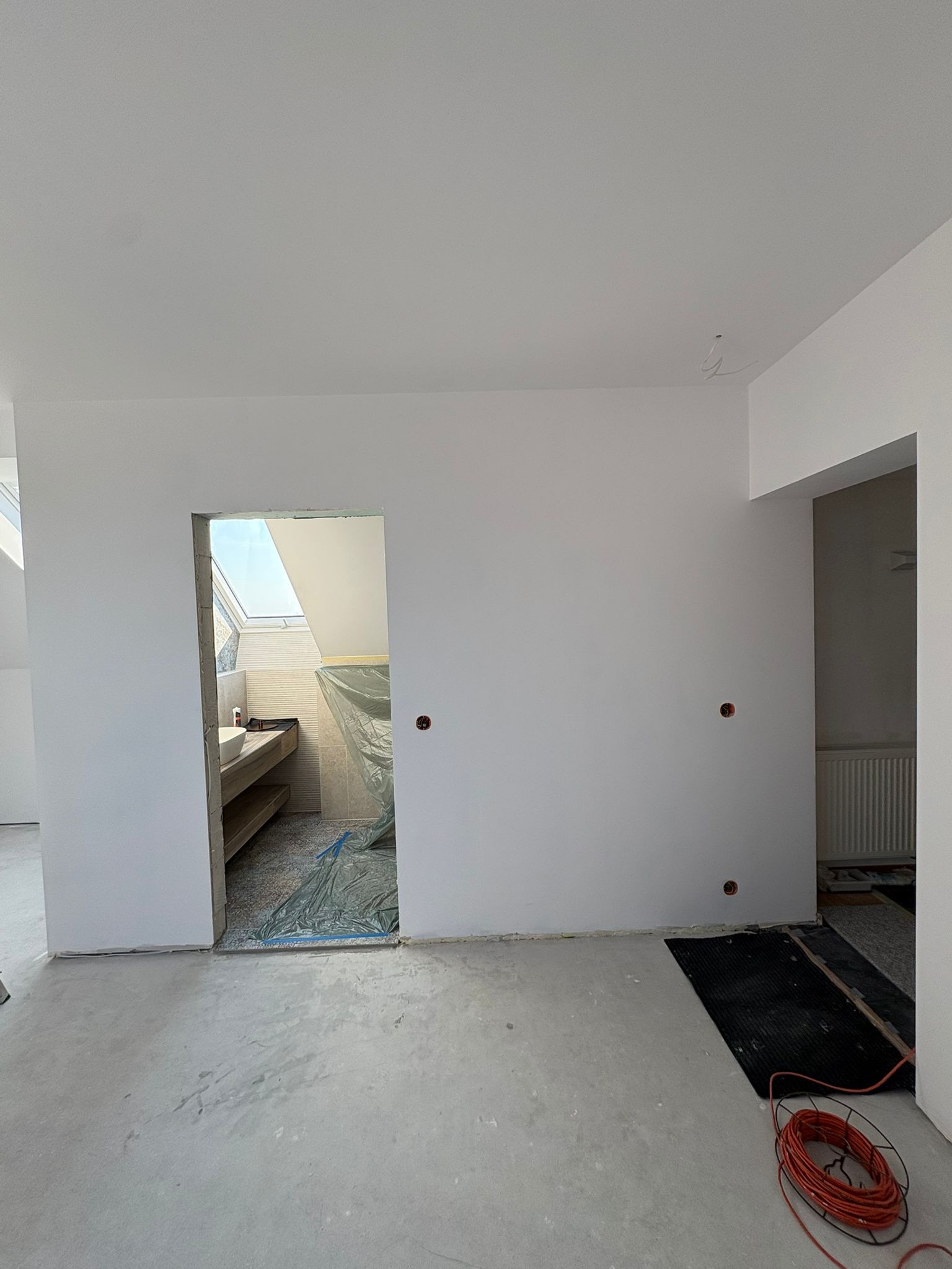 Wnętrze w trakcie prac wykończeniowych: białe ściany, surowa podłoga, widoczne instalacje elektryczne i fragment łazienki z oknem dachowym w tle.