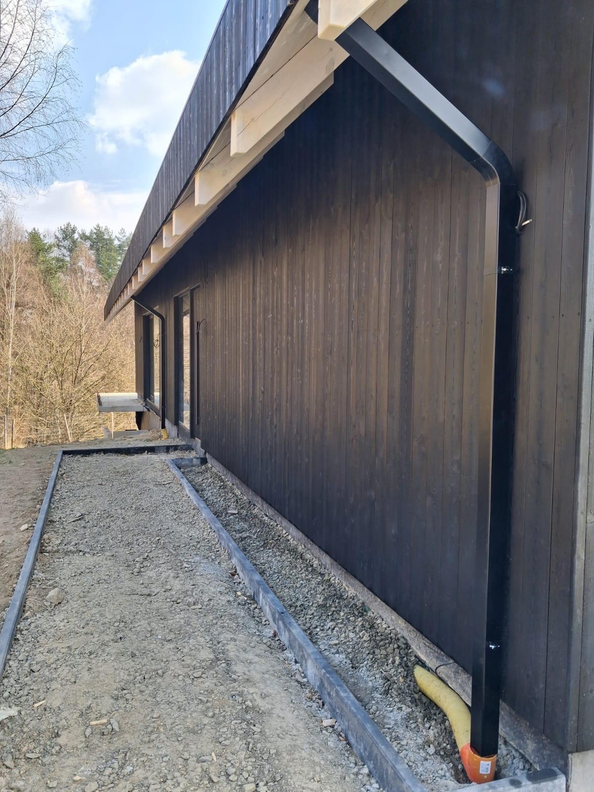 Elewacja wentylowana z ciemnego drewna, widok z boku, z czarną rynną i fragmentem terenu z kamieniami. Widoczne okna i elementy konstrukcyjne dachu.