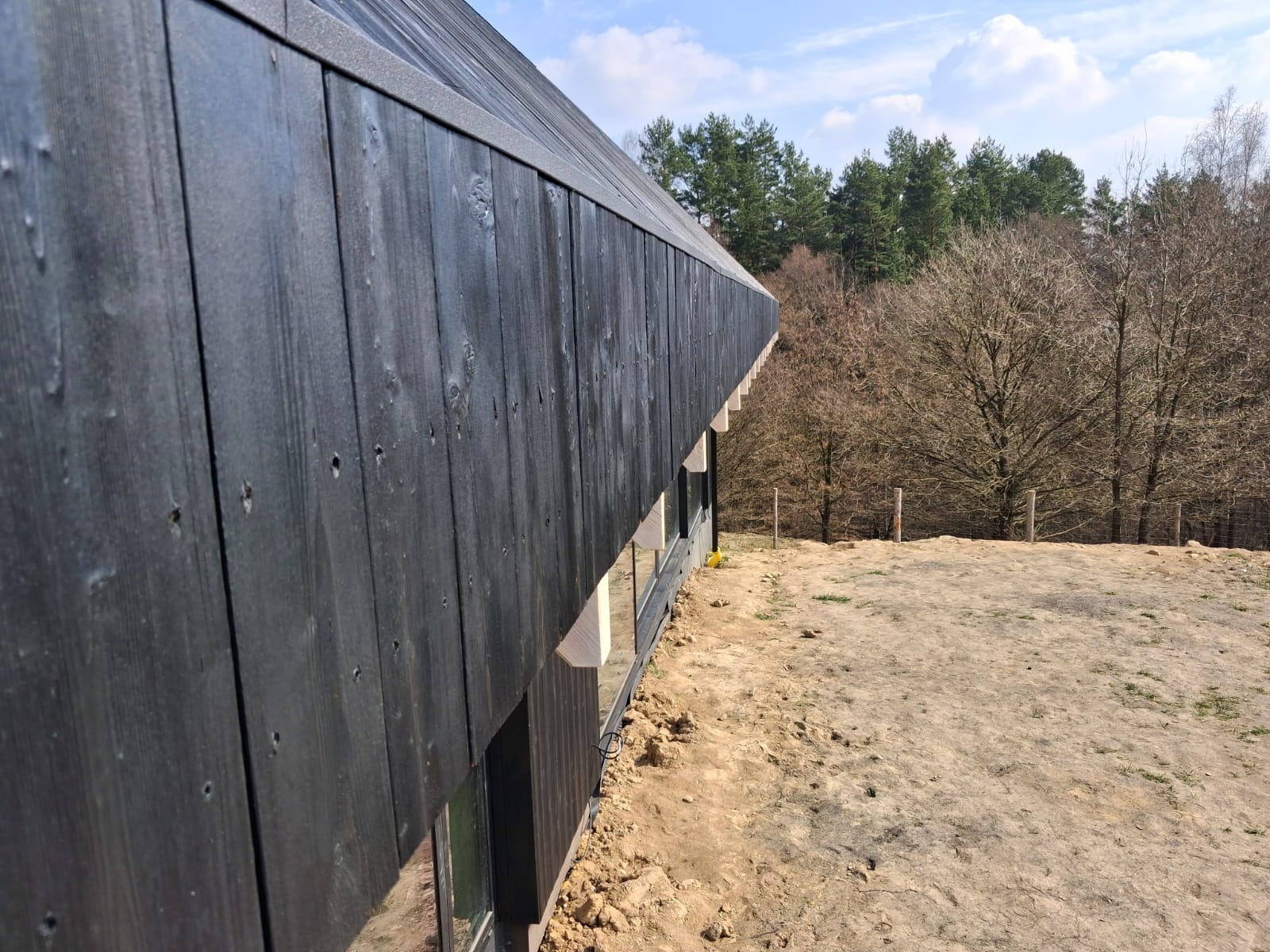 Czarna, drewniana elewacja wentylowana domu jednorodzinnego z widocznymi oknami i naturalnym otoczeniem leśnym w tle, uchwycona pod kątem.