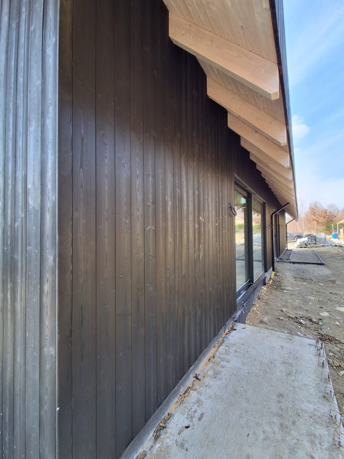 Ciemna, drewniana elewacja wentylowana domu jednorodzinnego z widocznymi oknami i fragmentem betonowego chodnika. Ujęcie z boku, w tle drzewa i błękitne niebo.