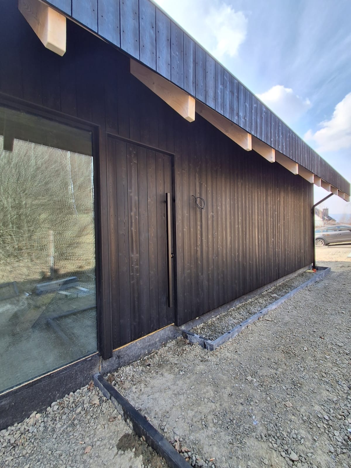 Czarna, drewniana elewacja wentylowana z widocznym oknem i drzwiami. Surowe wykończenie terenu wokół budynku. Ujęcie z dołu, podkreślające pionowe deski.