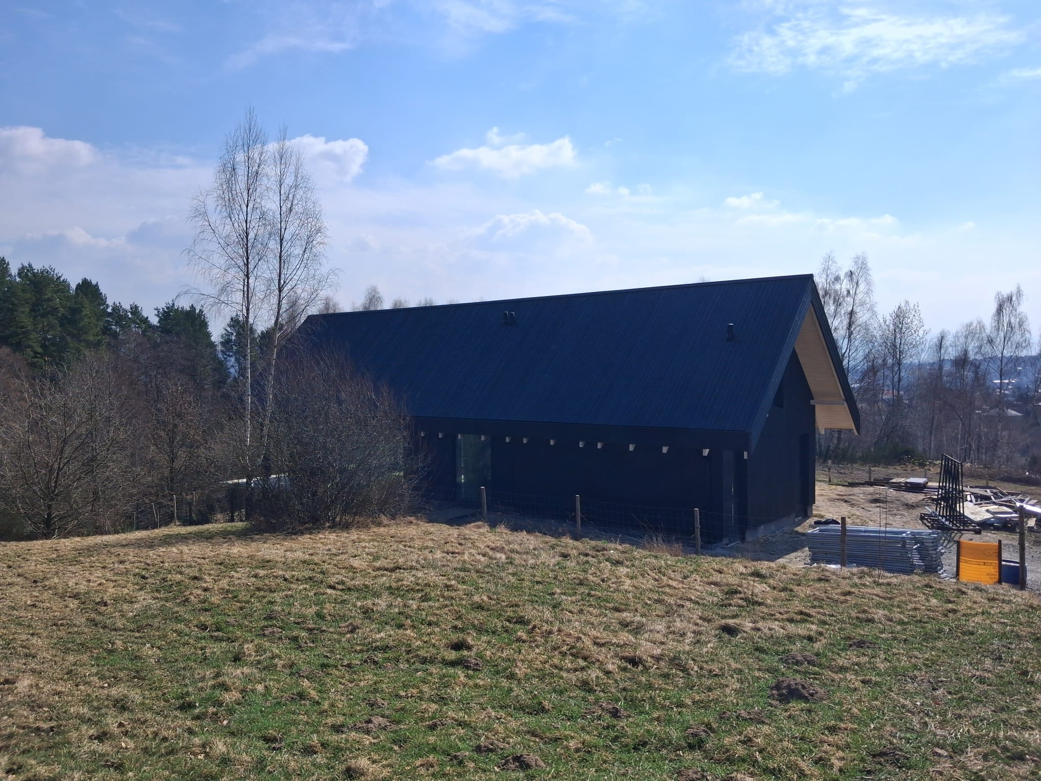 Nowoczesny dom modułowy z ciemną elewacją i dwuspadowym dachem, otoczony naturalnym krajobrazem z drzewami i łąką. Widoczne materiały budowlane obok domu.