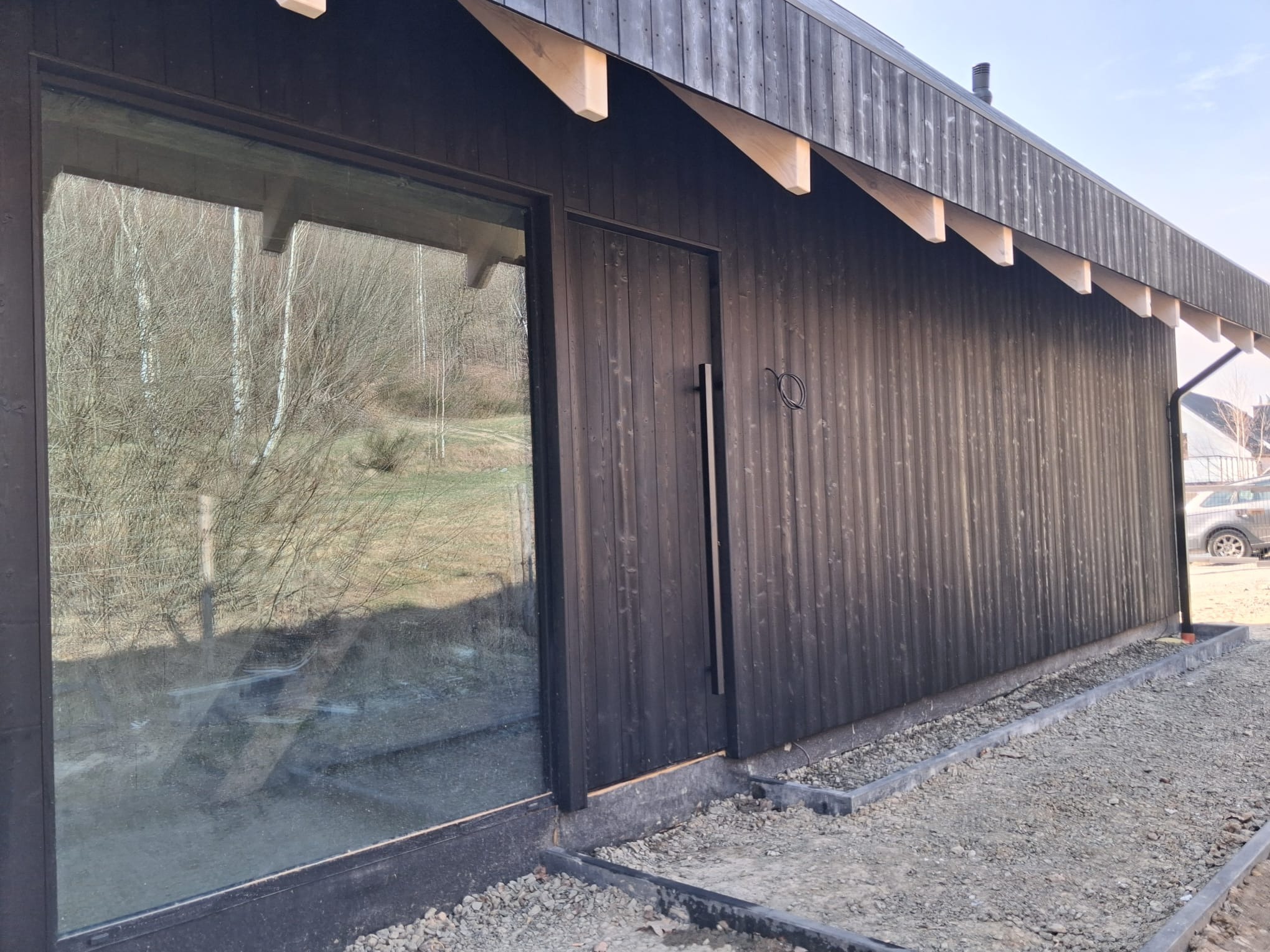 Nowoczesny dom modułowy z ciemną, drewnianą elewacją i dużym oknem, otoczony żwirem. Widoczne elementy konstrukcyjne dachu i minimalistyczne drzwi.