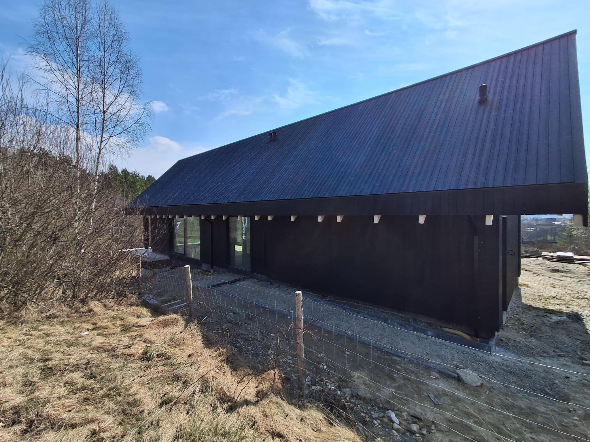 Czarny dom modułowy z blachy na dachu, widok z boku, ogrodzony siatką. Minimalistyczna architektura w surowym krajobrazie, błękitne niebo w tle.